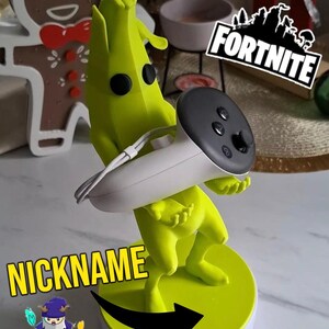 May include: A 3D printed yellow figurine of a character from the video game "Among Us" holding a gray video game controller. The figurine is standing on a white base with a yellow ring around the top. The text "NICKNAME" is written in black letters on the base. The text "FORTNITE" is written in white letters on a black background in the upper right corner of the image.