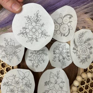 May include: Eight white fabric circles with black embroidery of different floral designs. The circles are arranged in a circular pattern on a woven surface.