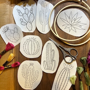 Op de afbeelding: Een set van acht borduurpatronen met verschillende soorten cactussen en vetplanten. De patronen zijn gedrukt op witte stof en klaar om te worden geborduurd. De patronen omvatten een tonvormige cactus, een stekelige peercactus, een saguaro-cactus en een verscheidenheid aan vetplanten.
