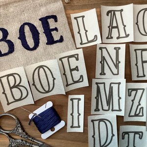 May include: A beige linen bag embroidered with the letters "BOE" in navy blue. Scattered around are individual letter decals in a serif font, along with a spool of navy thread, a needle, and vintage scissors. The letters are for crafting or personalization.