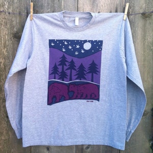 May include: A heather grey long-sleeve t-shirt with a purple and blue graphic of two bears in a forest with a full moon and stars. The graphic also includes the text "Silk Oak".