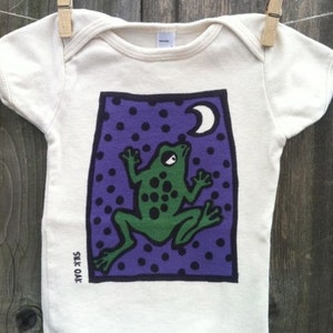 May include: A white baby onesie with a green frog looking up at a crescent moon on a purple background with white polka dots. The onesie has the text "Silk Oak" printed on the bottom left side.
