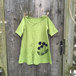 May include: A green linen tunic with a black floral print. The tunic has a scoop neck and short sleeves. It has a pocket on the left side.