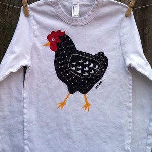 May include: White long-sleeved t-shirt with a black and white illustration of a chicken with a red comb. The text "Silk Oak" is printed on the bottom right of the design.