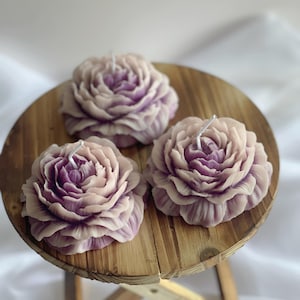 May include: Three purple and white floral candles on a wooden table. The candles are shaped like roses and have a delicate, layered design.
