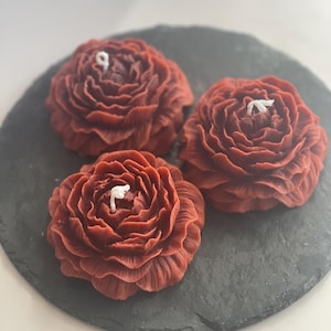 May include: Three red flower-shaped candles on a black slate surface. The candles have a white wick in the center.