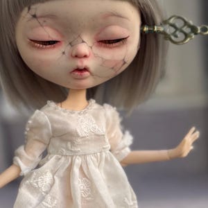 May include: A doll with short gray hair and a cracked face design featuring closed eyes and pink lips. The doll wears a white dress with embroidered floral patterns and pearl-like accents at the neckline. A metal key is tucked into the hair.