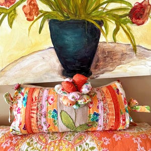 May include: A decorative pillow with a patchwork design in shades of orange, pink, and white. The pillow features a central burlap panel with a green leaf applique and is topped with fabric flower embellishments. The pillow rests on a patterned orange and white quilt.