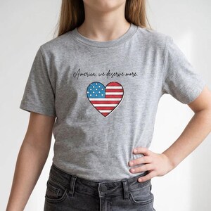 May include: A heather grey short-sleeved t-shirt featuring a heart-shaped graphic filled with the American flag pattern. Above the heart, the text "America, we deserve more." is printed in a black, cursive-style font on the chest area.