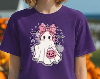 May include: A purple short-sleeve t-shirt featuring a graphic of a white ghost wearing a large pink polka-dot bow. The ghost holds a pink jack-o'-lantern bucket, surrounded by small bats, spiderwebs, and stars in a stylized illustration.