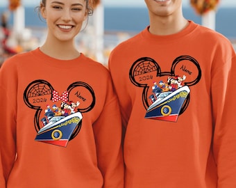 May include: Two orange crewneck sweatshirts featuring a black graphic of Mickey Mouse ears. Inside the ears is a cruise ship with cartoon characters, the text 2024, and a space for a name. The left sweatshirt includes a red polka dot bow.