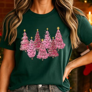 Pode incluir: Uma t-shirt de manga curta verde-escura com uma estampa gráfica de cinco árvores de Natal cor-de-rosa estilizadas. Cada árvore é decorada com linhas finas claras e rematada com uma pequena estrela amarela. A t-shirt tem gola redonda e bainha reta.