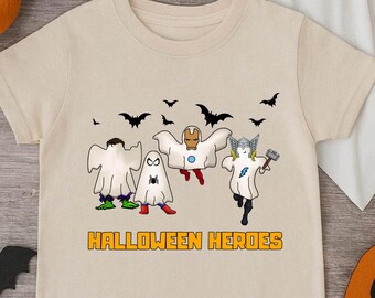 May include: A beige short-sleeve t-shirt featuring a graphic of four superheroes dressed as ghosts. The text HALLOWEEN HEROES is printed in orange below the characters, surrounded by small black bat silhouettes against a plain background.