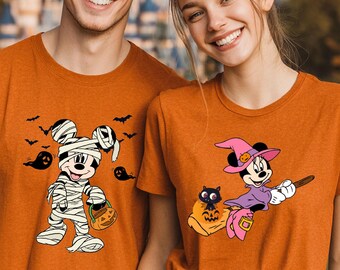 May include: Two orange short-sleeved t-shirts. The left shirt features a graphic of Mickey Mouse wrapped in bandages as a mummy holding a pumpkin bucket with black bats and ghosts. The right shirt shows Minnie Mouse as a witch on a broomstick.