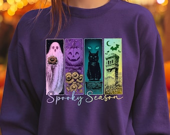 May include: A purple long-sleeve sweatshirt featuring a graphic print of four vertical panels depicting a ghost with a sunflower, a jack-o'-lantern with sunflowers, a black cat, and a tombstone with a bat, with the text Spooky Season below.