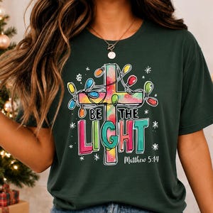 May include: A dark green t-shirt featuring a graphic of a watercolor-style cross wrapped in colorful Christmas lights. The text reads "BE THE LIGHT" in bold, multi-colored letters, with "Matthew 5:14" printed in white underneath the design.
