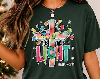 May include: A dark green t-shirt featuring a graphic of a watercolor-style cross wrapped in colorful Christmas lights. The text reads "BE THE LIGHT" in bold, multi-colored letters, with "Matthew 5:14" printed in white underneath the design.