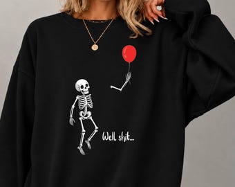 May include: A black crewneck sweatshirt featuring a white graphic of a skeleton with a detached arm holding a red balloon. The text "Well, shit..." is printed in white script below the skeleton. The sweatshirt has long sleeves and a ribbed collar.