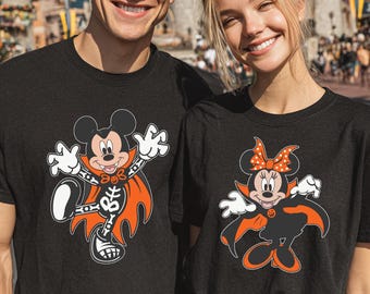 May include: Two black short-sleeved t-shirts featuring graphic prints. The left shirt displays Mickey Mouse dressed as a vampire with a skeleton motif. The right shirt features Minnie Mouse in a vampire costume with an orange polka dot bow.