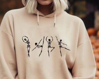 May include: A beige hooded sweatshirt featuring a graphic print of four black skeletons in various ballet poses across the chest. The garment includes a front kangaroo pocket, long sleeves with ribbed cuffs, and matching drawstrings at the neckline.