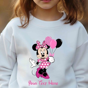 Puede incluir: Sudadera blanca con un gr&aacute;fico rosa de Minnie Mouse sosteniendo globos rosas y negros. El texto "Your Text Here" est&aacute; impreso debajo del gr&aacute;fico.