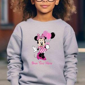 Puede incluir: Una sudadera gris con un dise&ntilde;o de Minnie Mouse rosa y blanco sosteniendo globos rosas y negros. El texto "Your Text Here" est&aacute; impreso debajo del dise&ntilde;o.