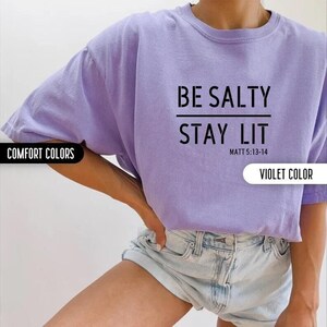 May include: A purple t-shirt with the text "BE SALTY STAY LIT" and the scripture reference "MATT 5:13-14" printed on the front. The shirt is made of a soft, comfortable material.
