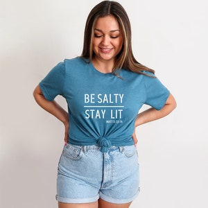 May include: A teal blue t-shirt with the text "BE SALTY STAY LIT" in white. The text "MATT 5:13-14" is printed in smaller white letters below the main text.