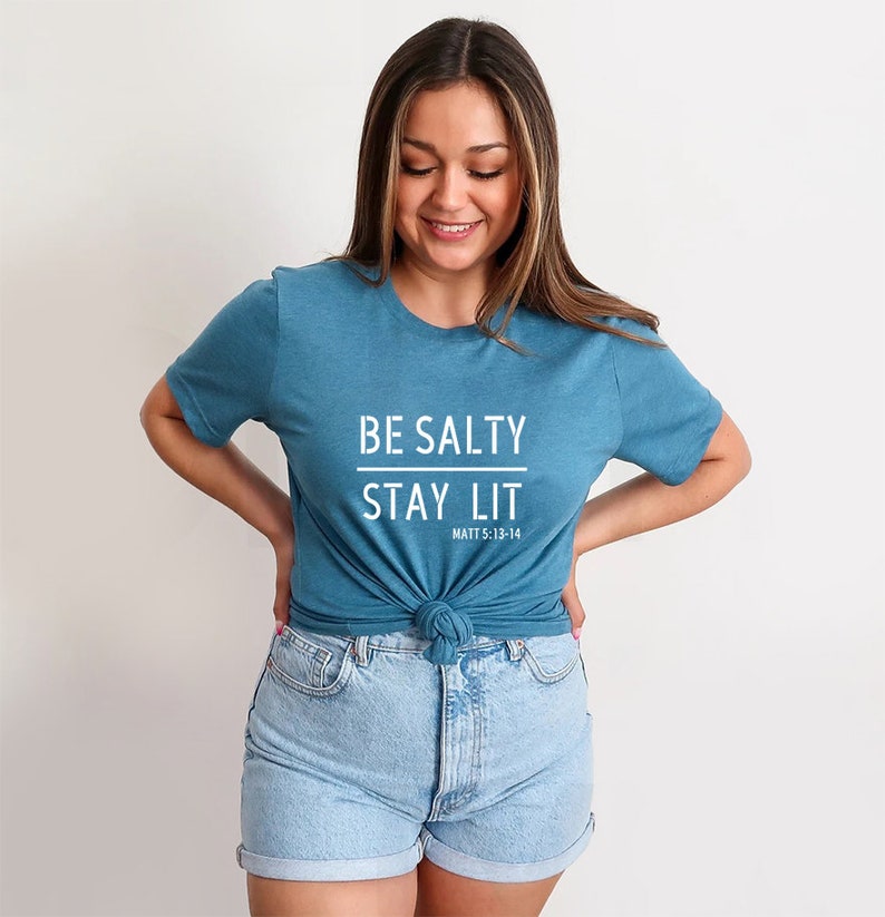 May include: A teal blue t-shirt with the text "BE SALTY STAY LIT" in white. The text "MATT 5:13-14" is printed in smaller white letters below the main text.