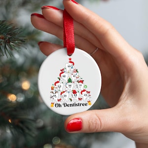 Puede incluir: Un adorno navide&ntilde;o blanco y redondo con una cinta roja. El adorno presenta un &aacute;rbol de Navidad de dibujos animados hecho de dientes con gorros de Pap&aacute; Noel. El texto "Oh Dentistree" est&aacute; debajo.