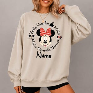 Pode incluir: Uma camisola bege de gola redonda com um gráfico da Minnie Mouse com um laço vermelho de bolinhas brancas. O texto diz Family Vacation 2020, Making Memories Together e Name. Pequenos ícones do Mickey e da Minnie estão nas laterais.