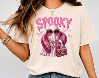 May include: A cream-colored t-shirt featuring a graphic of a ghost wearing pink sunglasses, a dark pink faux fur stole, and a matching handbag. The word SPOOKY is printed above the ghost in a dripping pink font, surrounded by small butterflies.