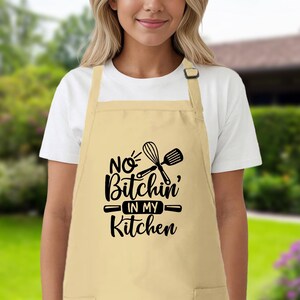 May include: A beige kitchen apron with black text that reads "No Bitchin' in my Kitchen" and a graphic of a whisk and rolling pin.