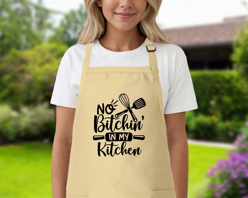 May include: A beige kitchen apron with black text that reads "No Bitchin' in my Kitchen" and a graphic of a whisk and rolling pin.