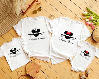 May include: White t-shirts and a onesie with a black airplane design featuring Mickey Mouse ears and the text "Disney Bound." One shirt has a red bow. The shirts are arranged on a wooden surface.