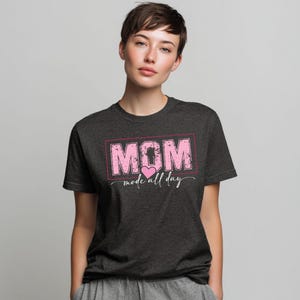 Puede incluir: Camiseta gris oscuro con la palabra "MOM" en rosa, dentro de un rect&aacute;ngulo rosa, con "mode all day" debajo. Un coraz&oacute;n rosa est&aacute; entre las palabras. La camiseta est&aacute; hecha de un material suave.
