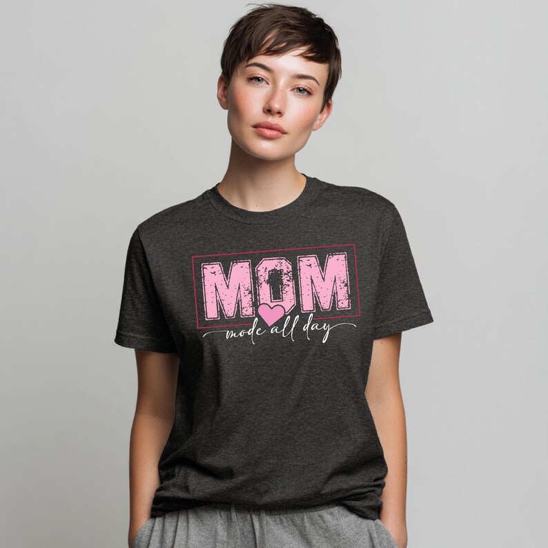 Puede incluir: Camiseta gris oscuro con la palabra "MOM" en rosa, dentro de un rect&aacute;ngulo rosa, con "mode all day" debajo. Un coraz&oacute;n rosa est&aacute; entre las palabras. La camiseta est&aacute; hecha de un material suave.