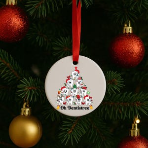 Puede incluir: Adorno navide&ntilde;o redondo y blanco con una cinta roja. El adorno presenta un dise&ntilde;o de &aacute;rbol de Navidad hecho de dientes de dibujos animados con gorros de Pap&aacute; Noel. El texto "Oh Dentistree" est&aacute; en la parte inferior del adorno.