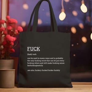 May include: Black tote bag with the word "FUCK" in white, along with a definition and related terms. The bag has two handles and is made of a sturdy material. The background includes red flowers in a pot and decorative lights.