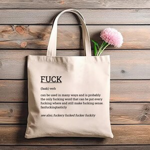 May include: Cream-colored tote bag with the word "FUCK" printed in black, along with a definition. The bag has a simple design with long handles. A pink flower with green stems is placed inside the bag.
