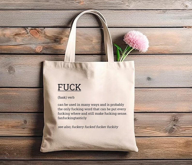 May include: Cream-colored tote bag with the word "FUCK" printed in black, along with a definition. The bag has a simple design with long handles. A pink flower with green stems is placed inside the bag.