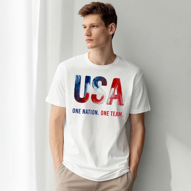 K&ouml;nnte beinhalten: Wei&szlig;es T-Shirt mit dem Wort "USA" in blau-roter Pinselstrich-Schrift. Darunter steht der Text "ONE NATION. ONE TEAM."