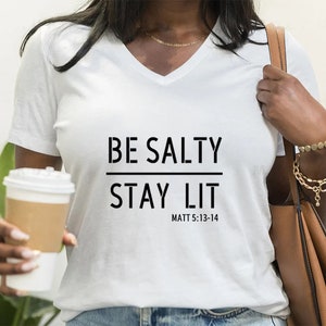 May include: White v-neck t-shirt with the text "BE SALTY STAY LIT" and "MATT 5:13-14" printed on the front.