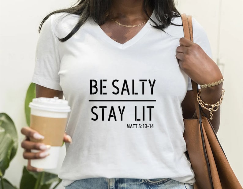 May include: White v-neck t-shirt with the text "BE SALTY STAY LIT" and "MATT 5:13-14" printed on the front.
