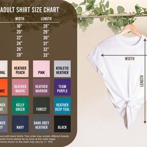 Puede incluir: Una camiseta blanca unisex colgada de una percha de madera, con una tabla de tallas a la izquierda. La tabla muestra tallas de camisa de S a 3X, con medidas de ancho y largo en pulgadas. Incluye muestras de color.