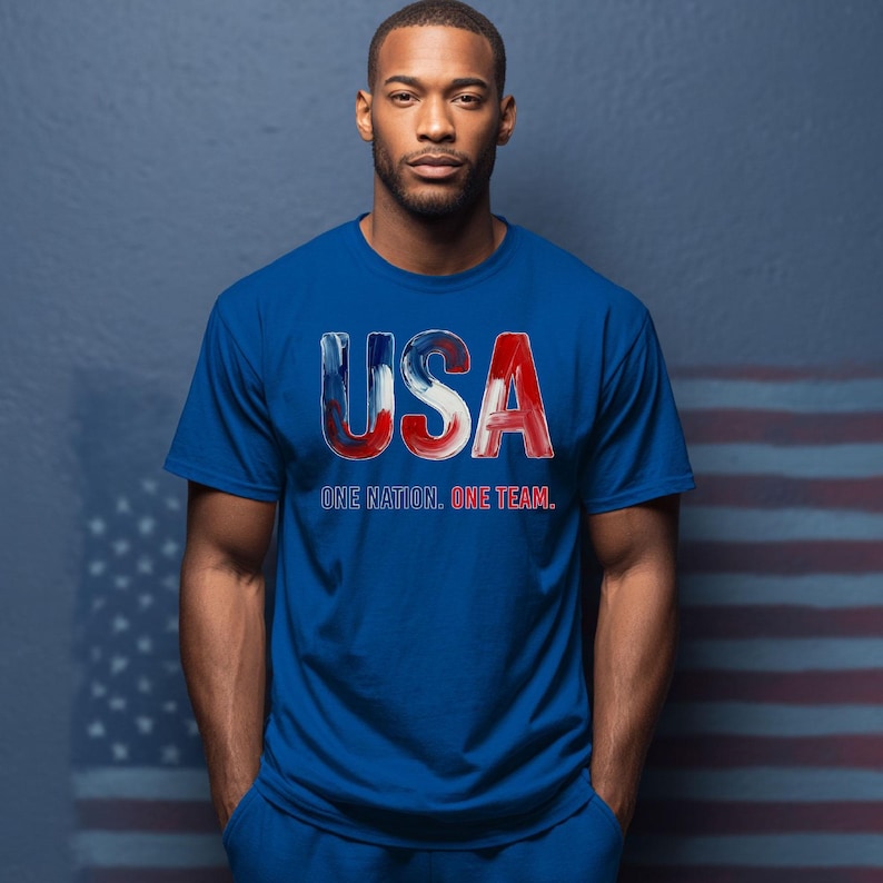 K&ouml;nnte beinhalten: Blaues T-Shirt mit dem Wort "USA" in Rot, Wei&szlig; und Blau und dem Text "ONE NATION. ONE TEAM." Das T-Shirt wird von einer Person getragen.