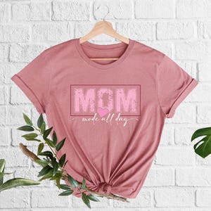 Puede incluir: Camiseta de color rosa polvoriento con la palabra "MOM" en un recuadro rosa, un coraz&oacute;n rosa y la frase "mode all day" en cursiva. La camiseta est&aacute; colgada de una percha de madera.