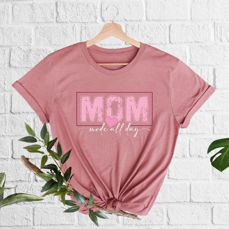 Puede incluir: Camiseta de color rosa polvoriento con la palabra "MOM" en un recuadro rosa, un coraz&oacute;n rosa y la frase "mode all day" en cursiva. La camiseta est&aacute; colgada de una percha de madera.