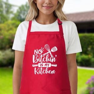 May include: Red kitchen apron with white text that says "No Bitchin' in my Kitchen" and a white whisk and spatula graphic.