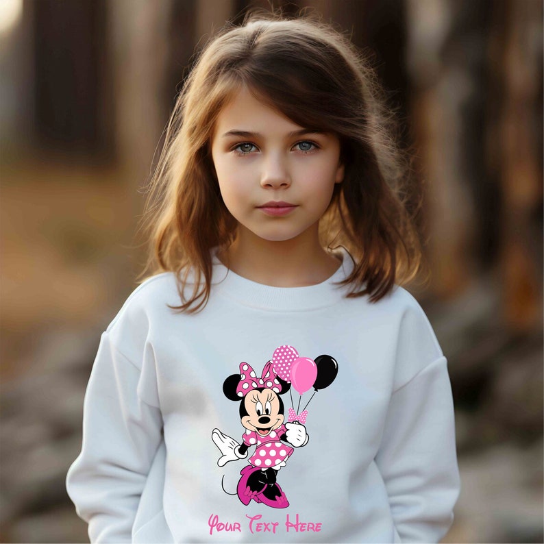 Puede incluir: Sudadera blanca con un gr&aacute;fico de Minnie Mouse rosa y negro sosteniendo globos rosas y negros. El texto "Your Text Here" est&aacute; impreso debajo del gr&aacute;fico.